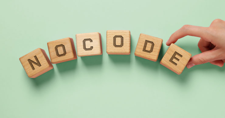 木製のキューブが並び、文字を形づくっている様子。手にした「E」のキューブを置くことで「NOCODE」という単語が完成する。少しの操作でDXが推進するイメージ。