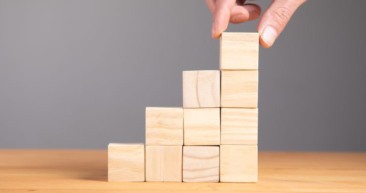 「JUST.DB」導入を段階的に進める「スモールスタート」の考え方を表すイメージ。木製のキューブを一つずつ積み上げ、着実な成長と成功へのステップを象徴している。