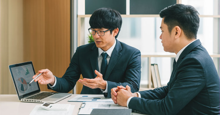 眼鏡の男性がノートPCの画面を示しながら、もう一人に説明しているビジネスマン2人の画像。「ノーコードツール導入」を成功させる伴走支援をイメージしたもの。